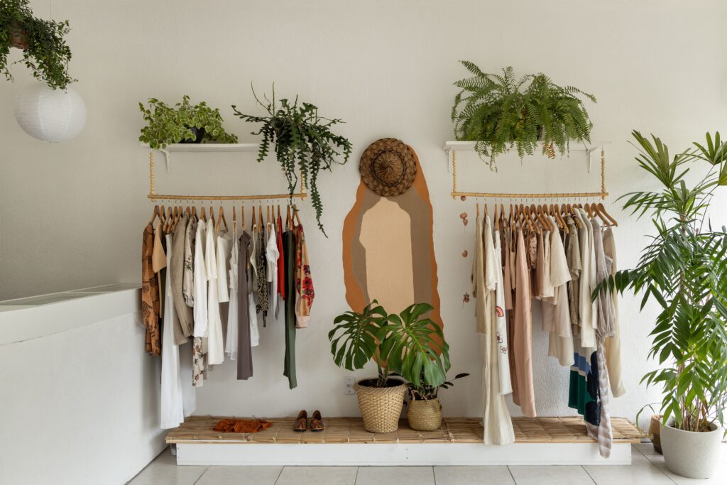 Interior de loja de moda circular estilo boho, com araras de madeira suspensas, decoração com plantas, pintura de arte orgânica na parede e exibição de roupas em tons terrosos.