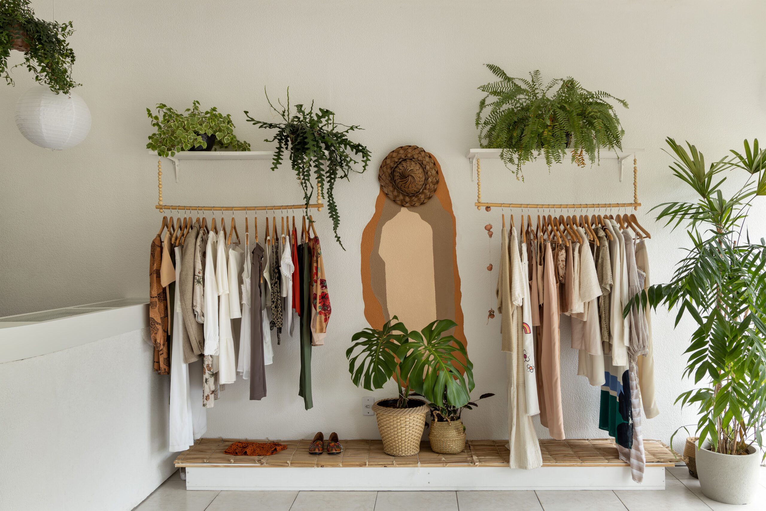 Interior de loja de moda circular estilo boho, com araras de madeira suspensas, decoração com plantas, pintura de arte orgânica na parede e exibição de roupas em tons terrosos.
