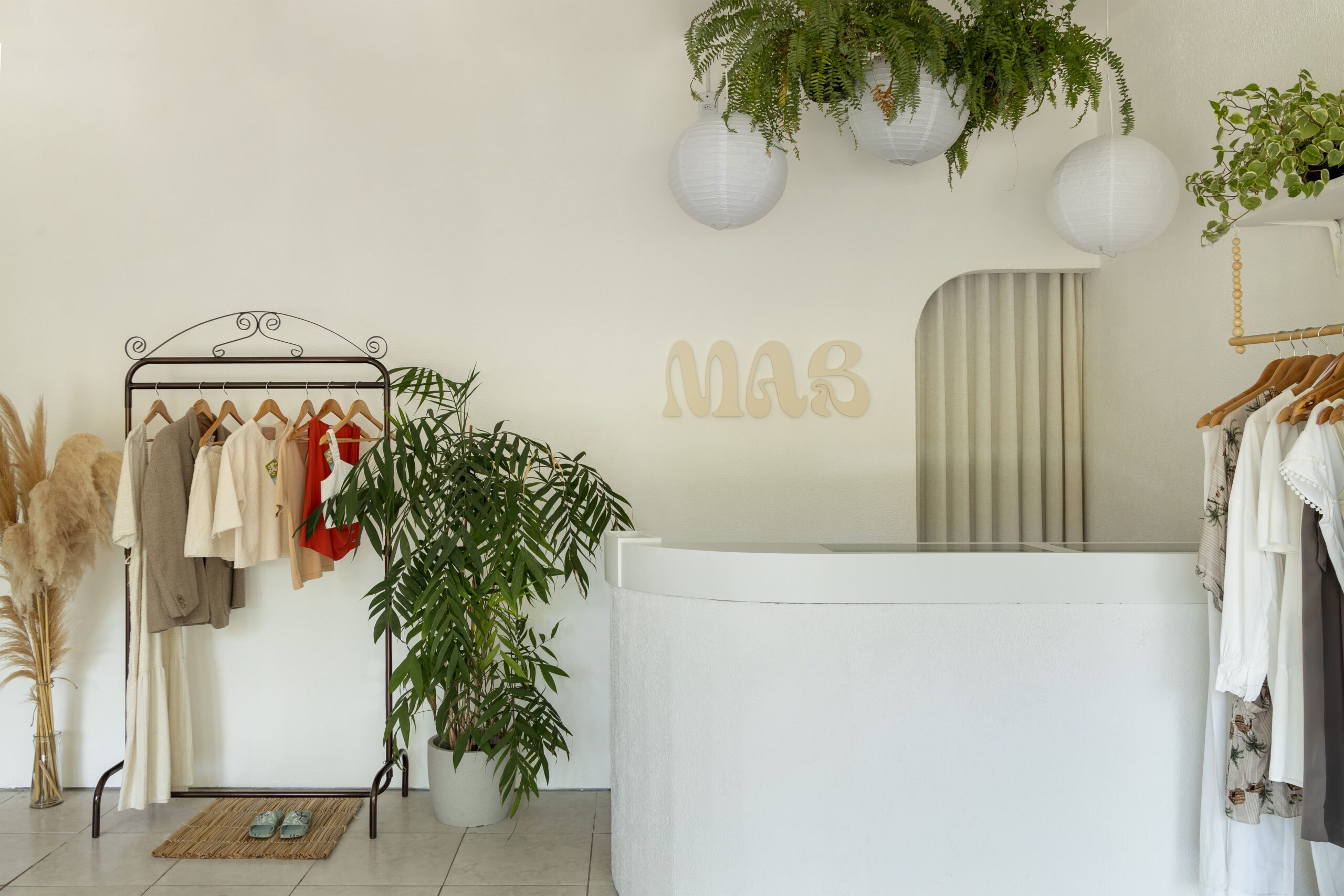 Balcão de atendimento curvo em gesso estilo boho da MAB Moda Circular, com logo da marca na parede, arara vintage e luminárias pendentes.