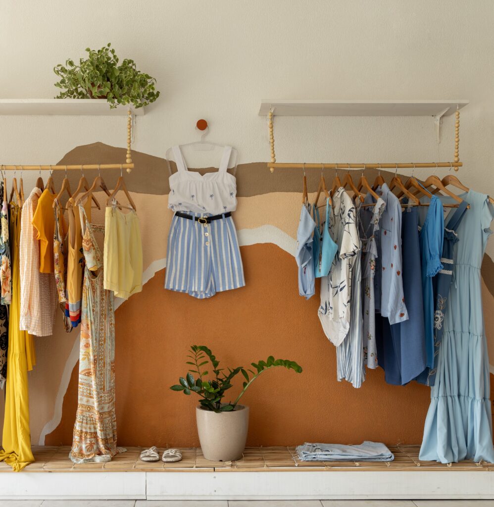Araras de madeira suspensas com roupas expostas em brechó estilo boho, com fundo de parede pintada com arte orgânica em tons terrosos e plantas decorativas.