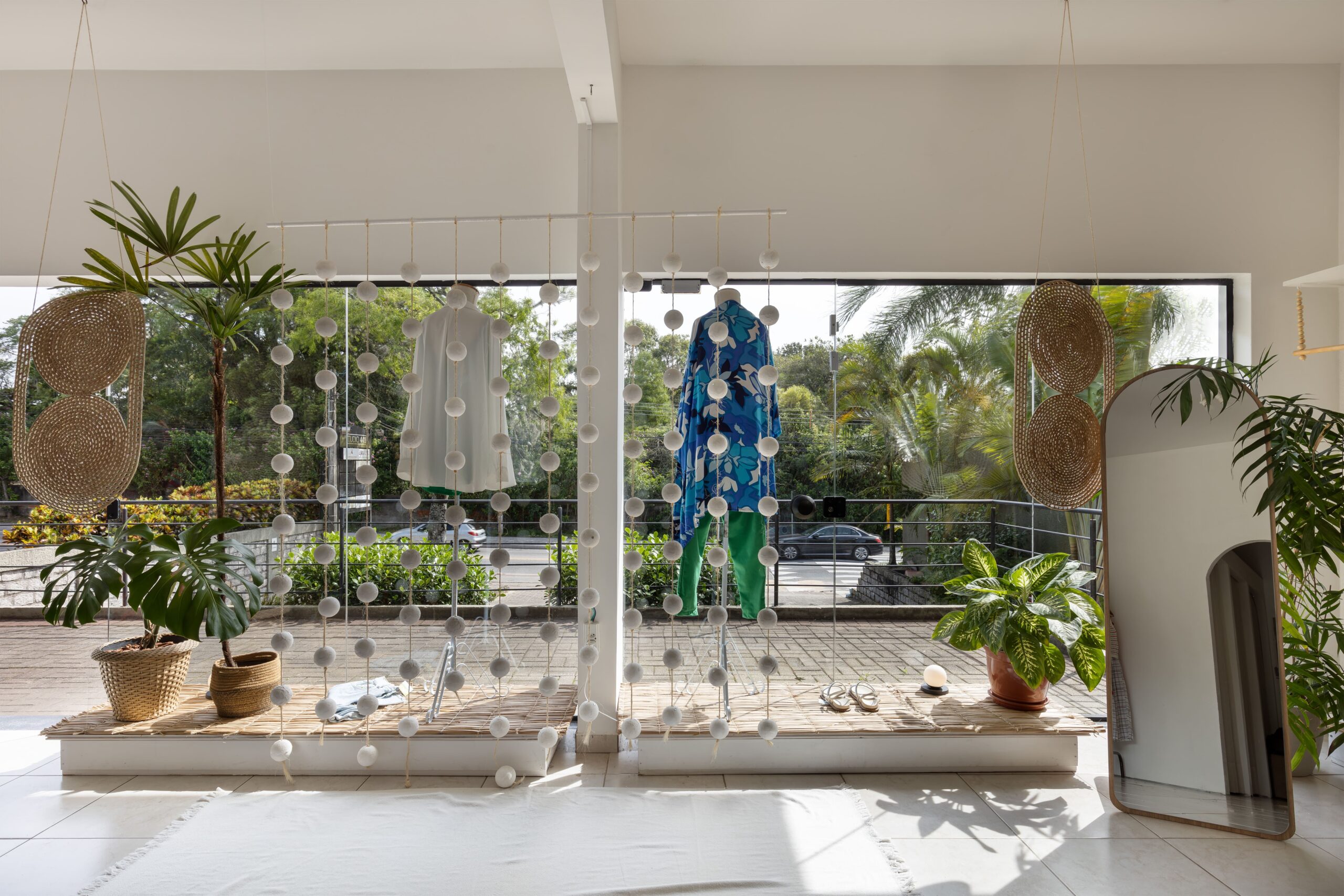 "Vitrine criativa de loja de roupas estilo boho, decorada com cortina de esferas de isopor em cordas de sisal, tablado de esteira de taboa e plantas, em Florianópolis.