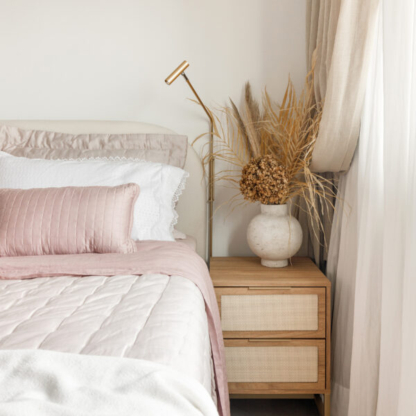 Cama de casal com enxoval rosa e branco, mesa de cabeceira de madeira com gavetas em palhinha, vaso com desidratados e arandela articulada dourada.