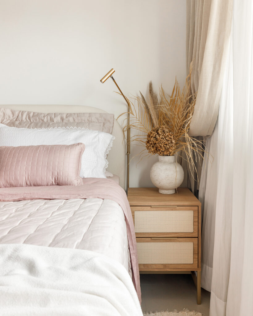 Cama de casal com enxoval rosa e branco, mesa de cabeceira de madeira com gavetas em palhinha, vaso com desidratados e arandela articulada dourada.