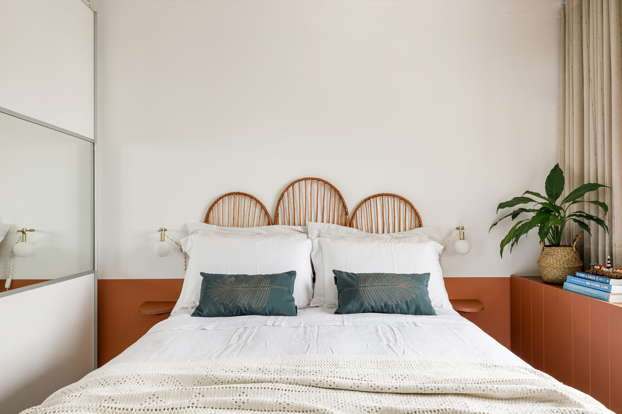 Cama de casal arrumada com enxoval branco, travesseiros verdes bordados, cabeceira tripla de bambu em arcos, pintura terracota em meia parede e arandelas com macramê.