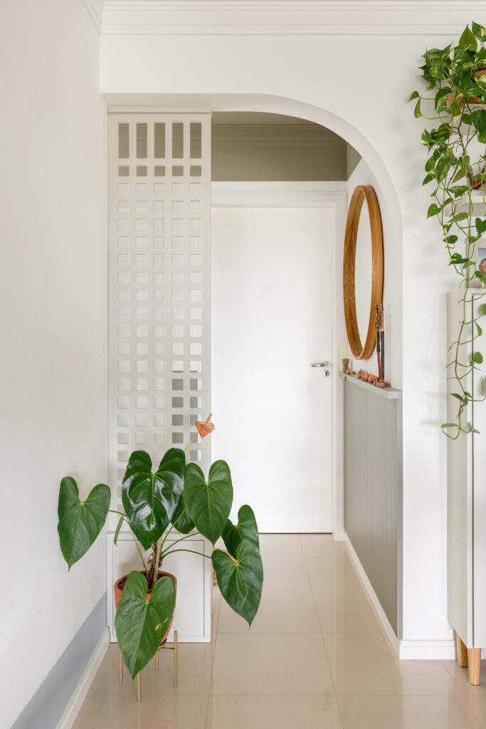 Passagem de hall de entrada para sala de estar com pórtico em arco, divisor de ambientes muxarabi branco, planta antúrio e espelho redondo.