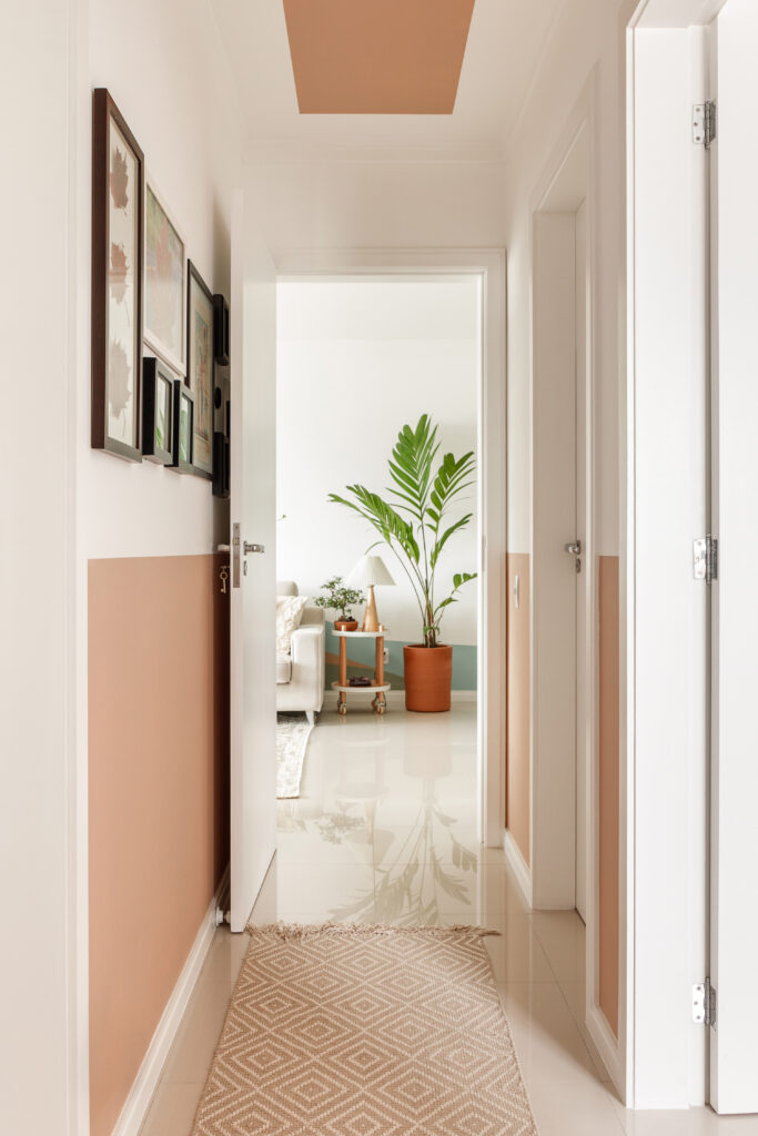 Corredor de apartamento com pintura criativa em meia parede e detalhe no teto em tom terroso, composição de quadros e passarela geométrica.