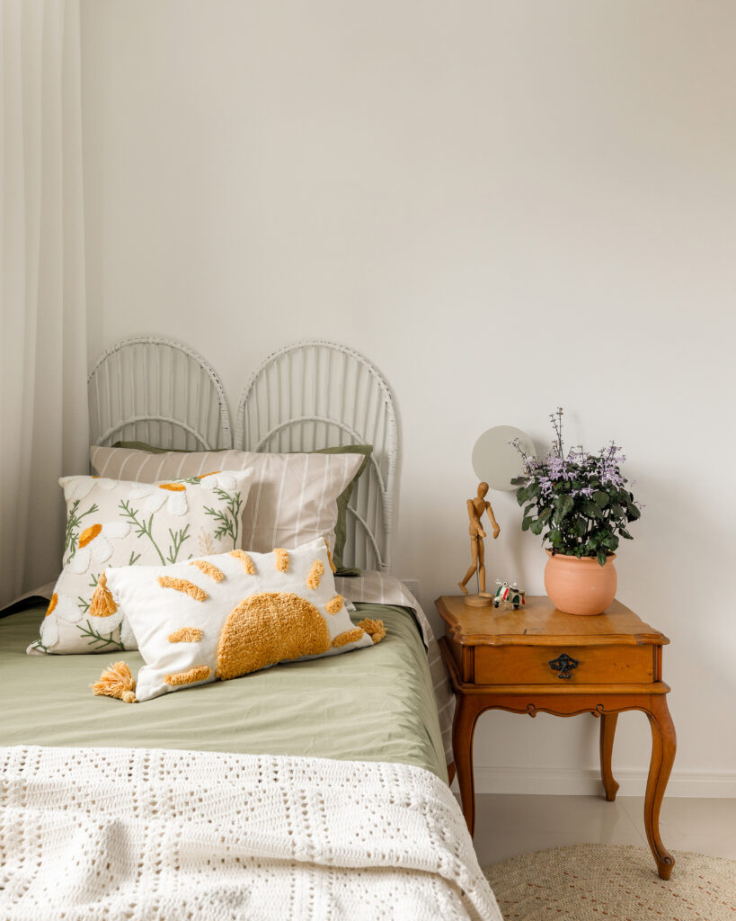 Quarto de solteiro decorado no estilo Boho Chic com cabeceira de vime branca, almofadas bordadas de sol e flores, mesa de cabeceira vintage de madeira e roupa de cama verde.
