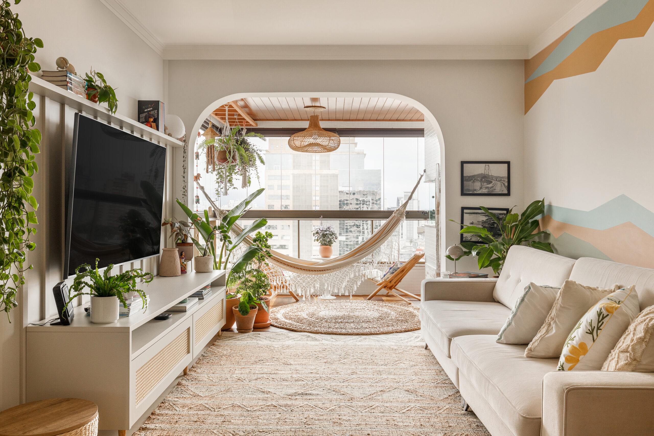 Sala de estar integrada com sacada no estilo Boho e Farmhouse, com sofá claro, rack branco com palhinha, plantas e rede de descanso em Florianópolis.