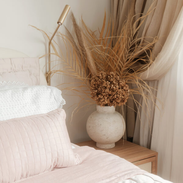 Cama de casal com enxoval rosa e branco, mesa de cabeceira de madeira com gavetas em palhinha, vaso com desidratados e arandela articulada dourada.