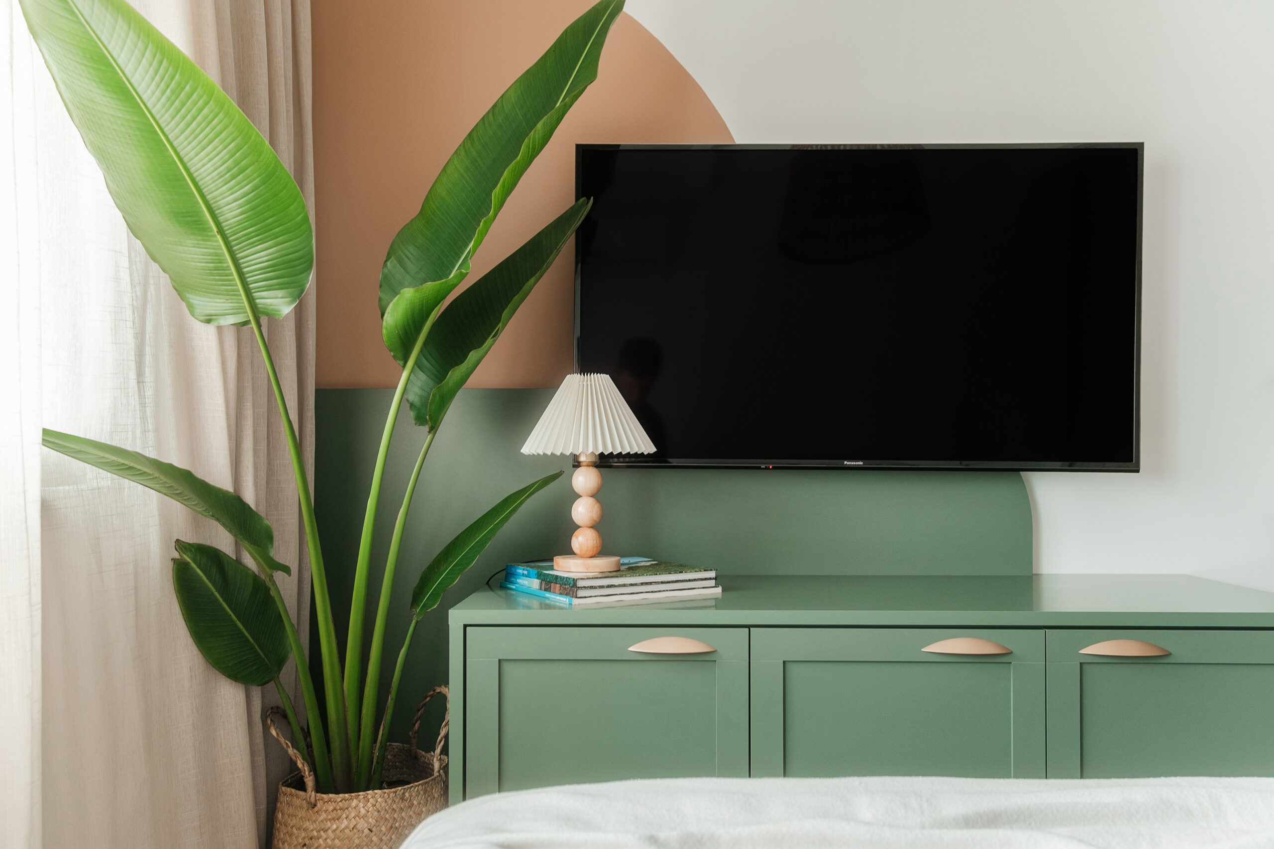 Parede da TV na suíte master com pintura artística geométrica em tons terracota e verde, móvel aparador verde com puxadores de concha, luminária plissada e planta Ravenala em cesto de palha.