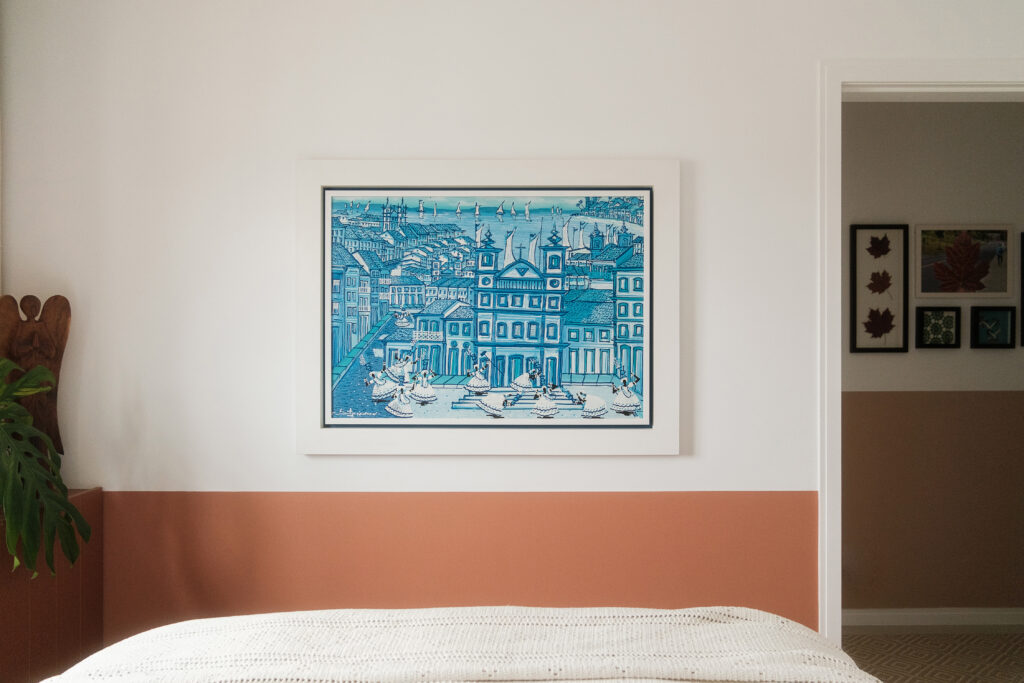 Quarto de hóspedes de casal com pintura em meia parede na cor terracota, cama feita e quadro com gravura em tons de azul retratando a cultura açoriana.