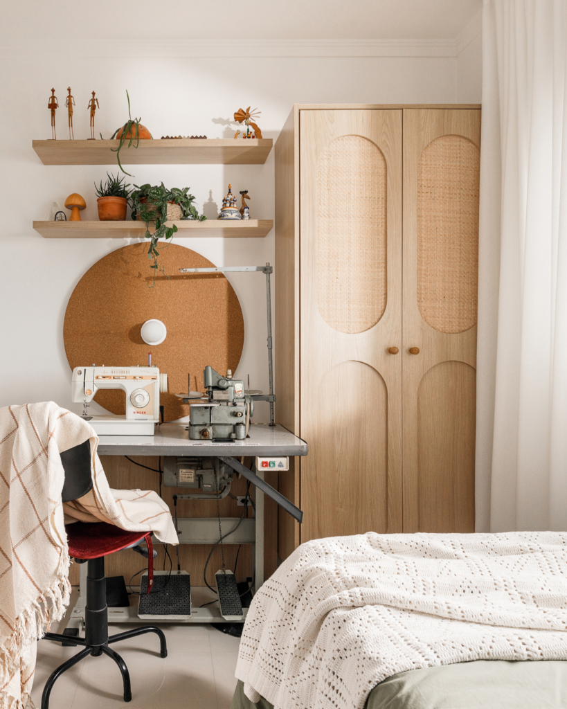 Ateliê de costura integrado ao quarto de solteiro com bancada de máquina de costura, painel redondo de cortiça, prateleiras e armário amadeirado com palhinha.
