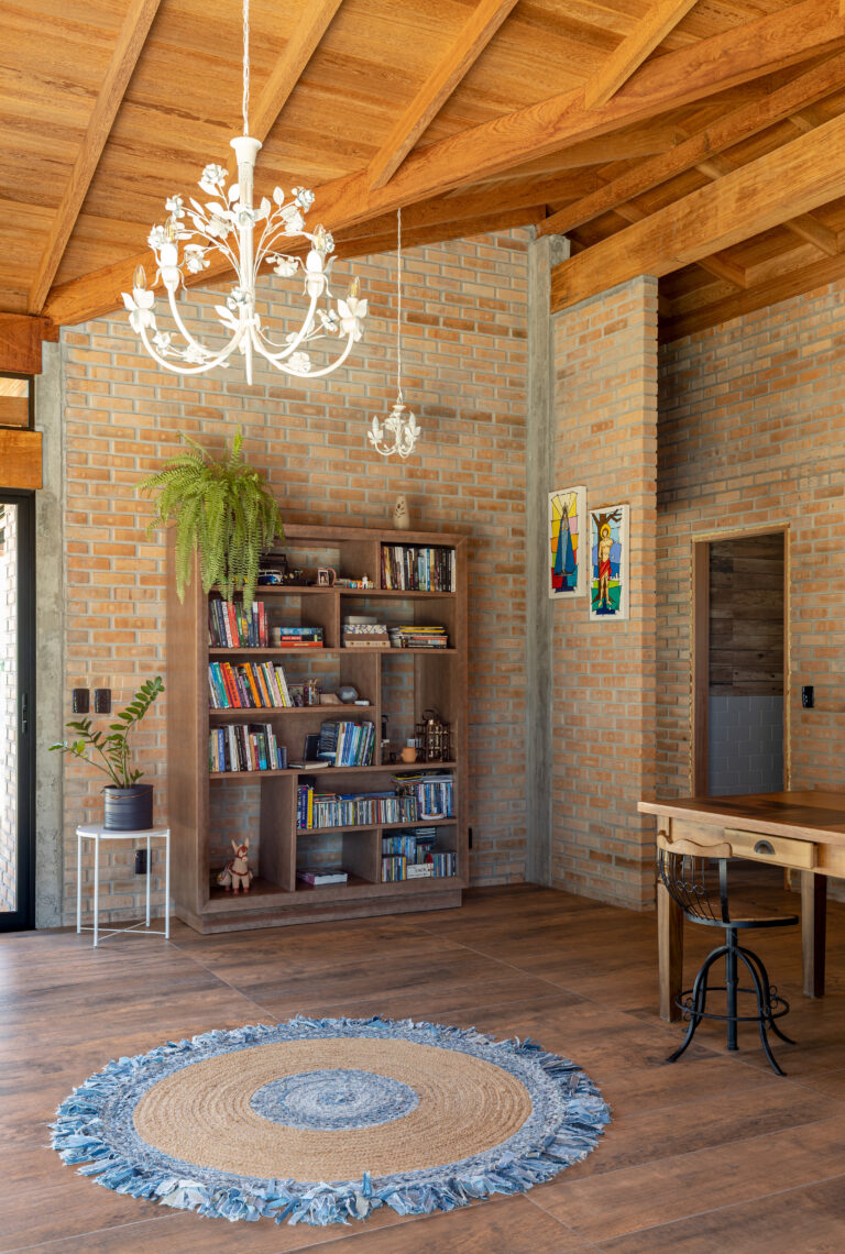 Sala de estar com estante de livros em madeira, parede de tijolo maciço aparente, teto com vigas de madeira inclinadas e lustre clássico, Casa Lar do Samba