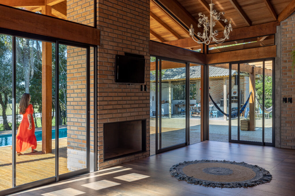 Sala de estar com lareira em tijolo maciço aparente, esquadrias amplas de vidro integradas ao deck, piscina e área gourmet externa, Casa Lar do Samba