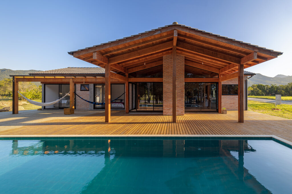 Vista frontal da piscina com deck de madeira e varanda com forro e vigas de madeira aparente, Casa Lar do Samba